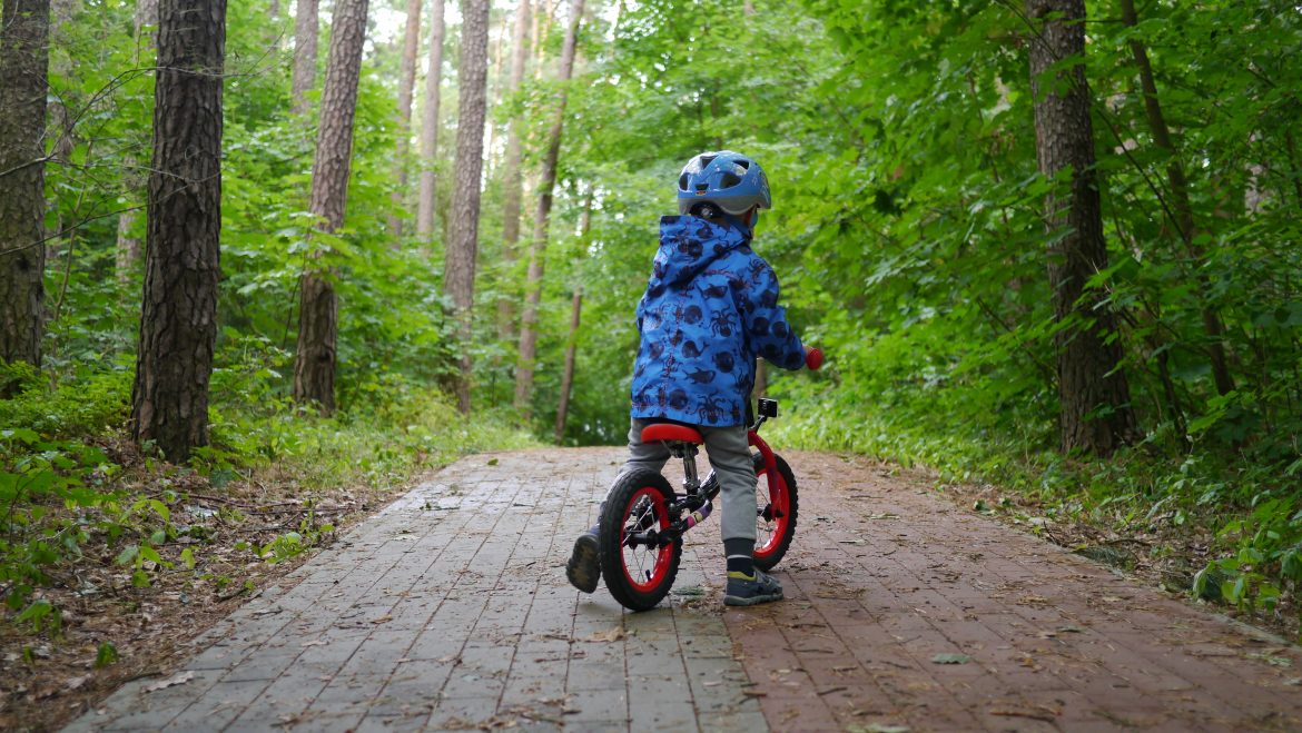 Dziecko w kasku jedzie na rowerze na drodze w lesie wśród drzew.